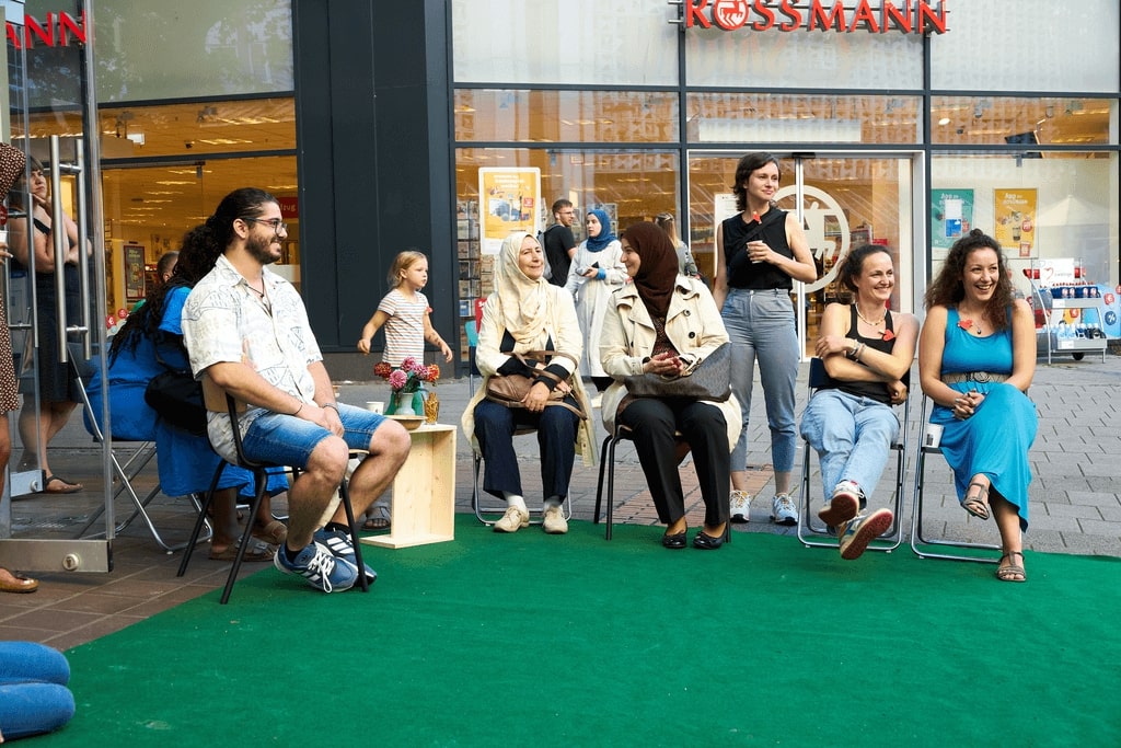 Menschen sitzen auf Klappstühlen auf einer Kunstrasenfläche vor dem UMZU-Laden im Hanseatenhof.
