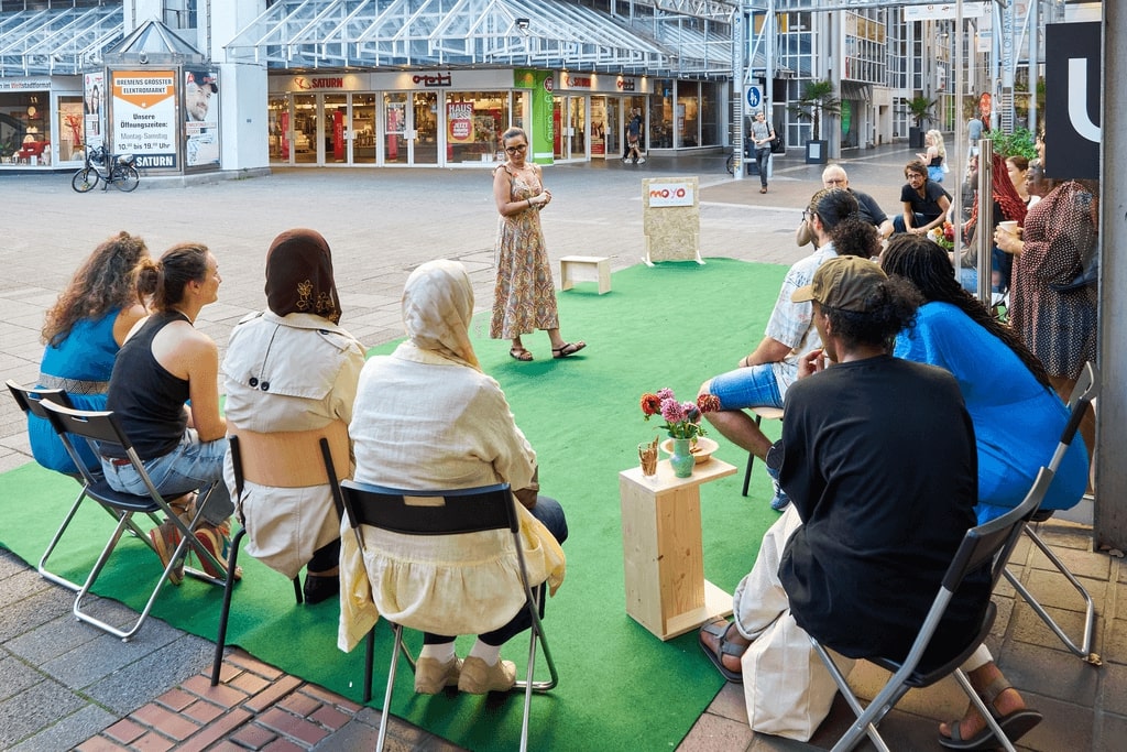 Menschen sitzen auf Klappstühlen auf einer Kunstrasenfläche vor dem UMZU-Laden im Hanseatenhof und hören einer Rednerin zu.