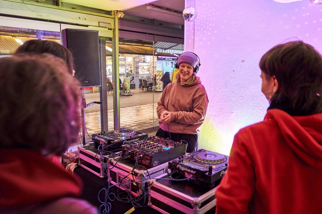 Blick auf ein DJ-Pult, hinter dem eine junge Frau steht und lacht.