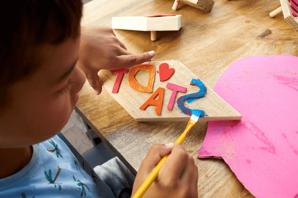 Kind bemalt Buchstaben auf einem Holzstück, welche zusammen das Wort Stadt ergeben.